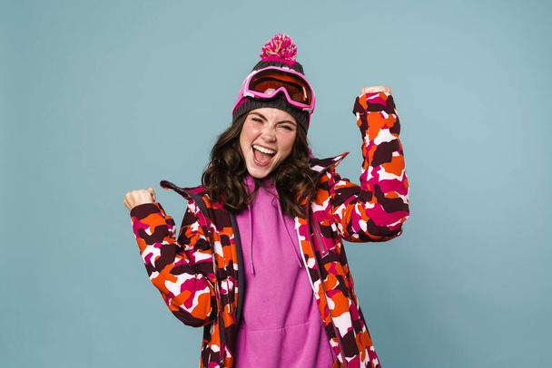 Excited young woman wearing snowboard jacket celebrating isolated over blue background - Foto, Imagen