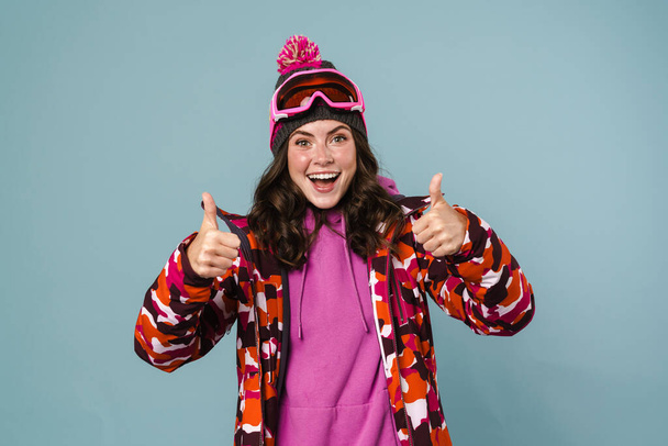Happy young woman wearing snowboarding gear standing isolated over blue background - Zdjęcie, obraz
