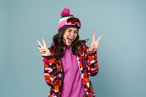 Happy young woman wearing snowboarding gear standing isolated over blue background - Foto, Imagen