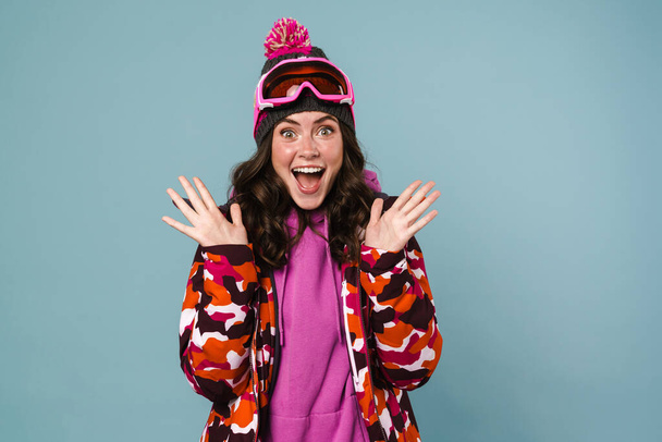 Happy excited young woman wearing snowboarding gear standing isolated over blue background - Fotó, kép