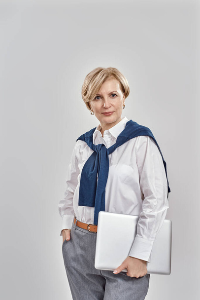 Портрет елегантної жінки середнього віку в діловому одязі, що тримає ноутбук, дивлячись на камеру, позуючи ізольовано на сірому фоні
 - Фото, зображення