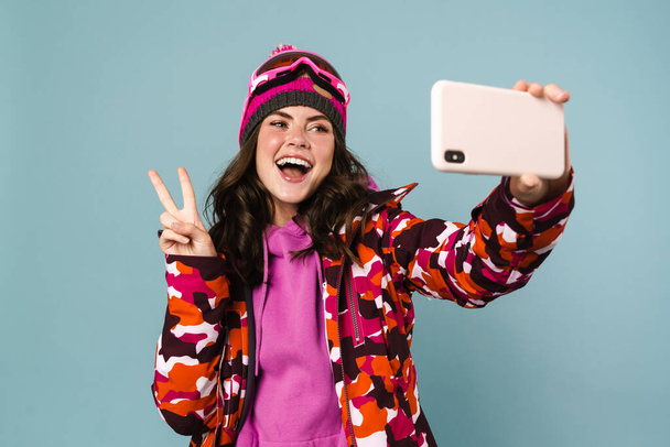 Wesoła młoda kobieta ubrana w sprzęt snowboardowy robi sobie selfie na niebieskim grzbiecie - Zdjęcie, obraz