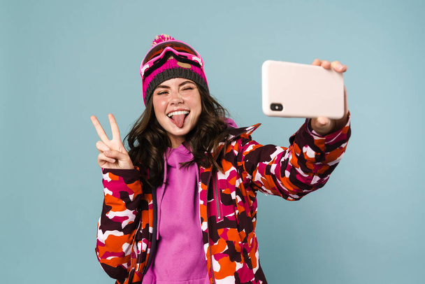 Cheerful young woman wearing snowboard gear taking a selfie isolated over blue backgorund - Foto, Bild