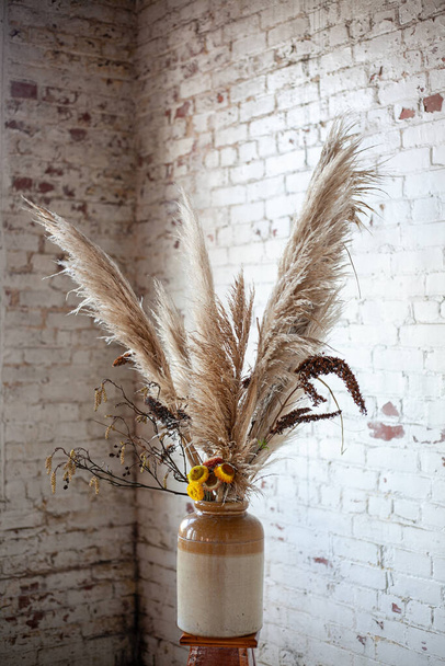 dry flowers and fluffy reeds in a beige ceramic vase on a wooden stool against a white brick wall background - Фото, изображение