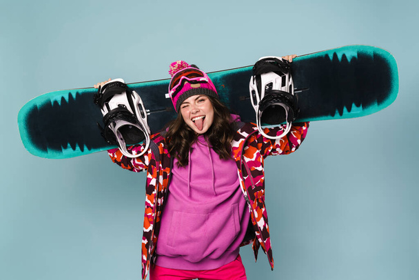 Happy sportswoman posing with snowboard isolated over blue wall - Foto, afbeelding
