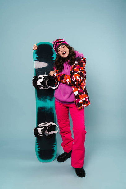 Happy sportswoman posing with snowboard isolated over blue wall, full length - Fotó, kép