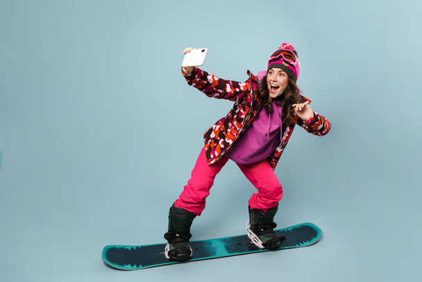 Cheerful sportswoman taking selfie on cellphone while posing on snowboard isolated over blue wall - Фото, зображення