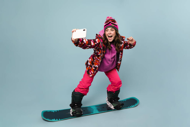 Cheerful sportswoman taking selfie on cellphone while posing on snowboard isolated over blue wall - Zdjęcie, obraz