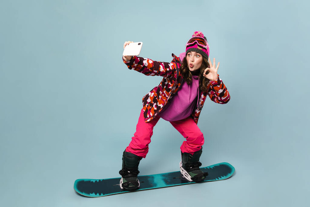 Cheerful sportswoman taking selfie on cellphone while posing on snowboard isolated over blue wall - 写真・画像