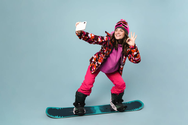 Cheerful sportswoman taking selfie on cellphone while posing on snowboard isolated over blue wall - Zdjęcie, obraz