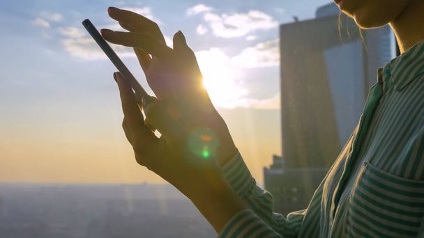 Nő használ okostelefon ellen Sunset cityscape keresztül ablak felhőkarcoló - Fotó, kép