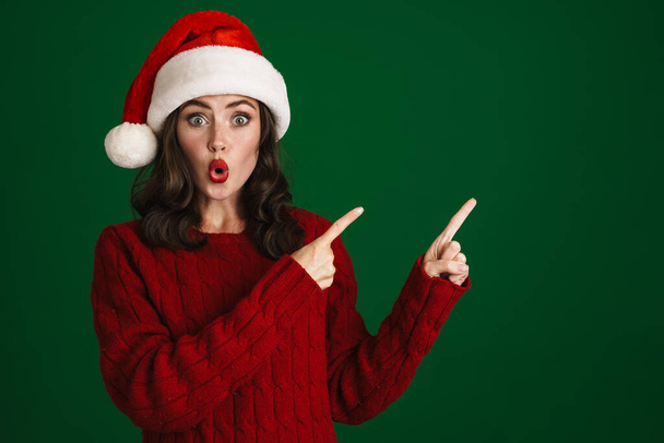 Beautiful excited young woman in red sweater and hat over green background, presenting copy space - Фото, изображение