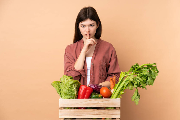 Молодая фермерская девочка со свежесобранными овощами в коробке показывает знак молчания жест положить палец в рот - Фото, изображение