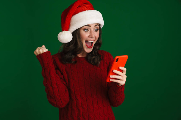 Nice excited girl in Santa Claus hat making winner gesture and using cellphone isolated over green background - Foto, afbeelding