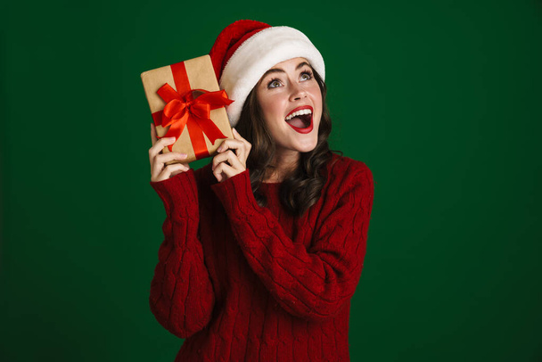 Charming excited girl in Santa Claus hat posing with Christmas gift isolated over green background - Foto, imagen