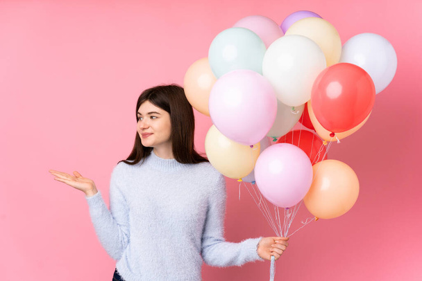 Young Ukrainian teenager girl holding lots of balloons over isolated pink background holding copyspace imaginary on the palm - Photo, Image