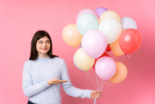 Young Ukrainian teenager girl holding lots of balloons over isolated pink background extending hands to the side for inviting to come - Photo, Image