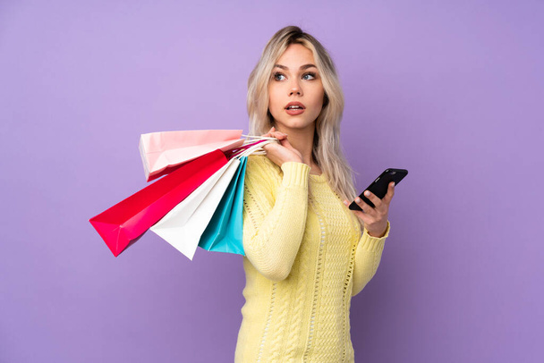 Teenager blonde Mädchen über isoliertem lila Hintergrund mit Einkaufstaschen und einem Mobiltelefon - Foto, Bild