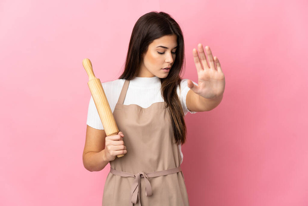 Teenager Brazilian girl holding a rolling pin isolated on pink background making stop gesture and disappointed - Фото, зображення