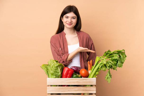 Молодая фермерская девушка со свежесобранными овощами в коробке представляет идею, глядя улыбаясь в сторону - Фото, изображение