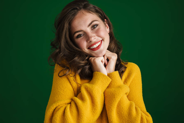 Cheerful beautiful young woman in sweater standing isolated over green background, looking at camera - Foto, Bild