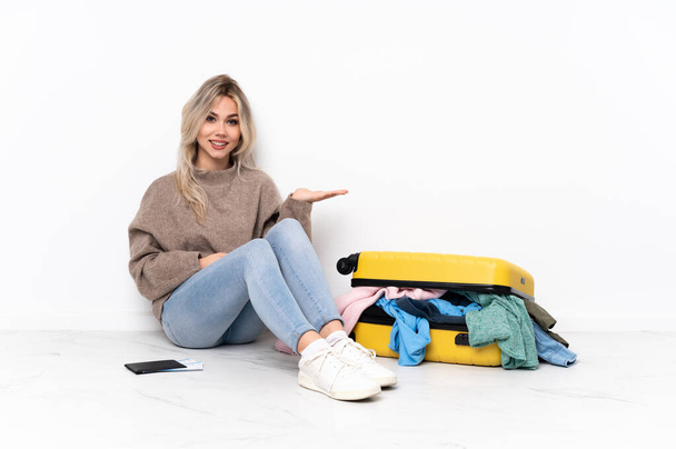 Adolescente ragazza bionda con una valigia piena di vestiti seduta sul pavimento che presenta un'idea mentre guarda sorridente verso - Foto, immagini