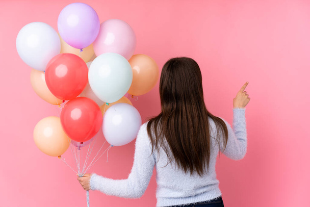 Молода українська дівчина-підліток тримає багато повітряних кульок над ізольованим рожевим фоном, вказуючи назад вказівним пальцем. - Фото, зображення
