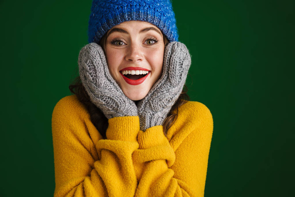 Cheerful beautiful young woman in sweater and hat standing isolated over green background, posing in mittens - Foto, afbeelding