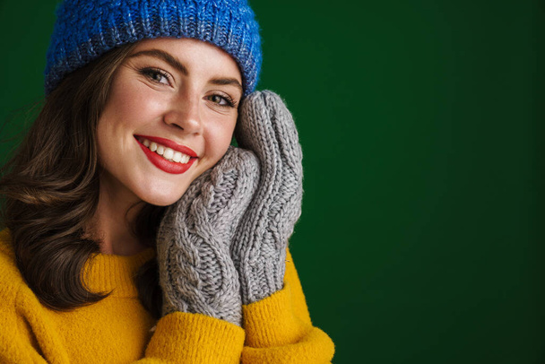 Beautiful happy girl in knit hat and mittens smiling at camera isolated over green background - Fotografie, Obrázek