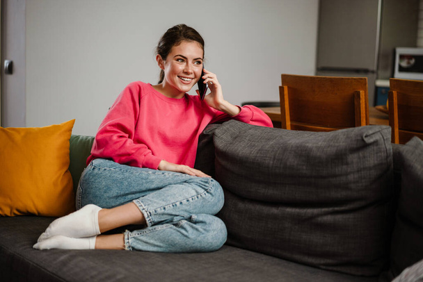 Beautiful joyful girl talking on cellphone and smiling while sitting on couch at home - Zdjęcie, obraz