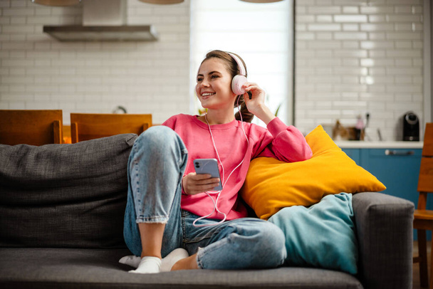 Nice joyful girl listening music with headphones and cellphone while sitting on couch at home - Valokuva, kuva