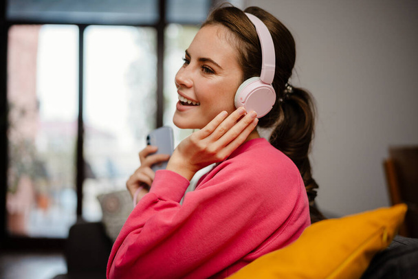 Nice joyful girl listening music with headphones and cellphone while sitting on couch at home - Foto, afbeelding