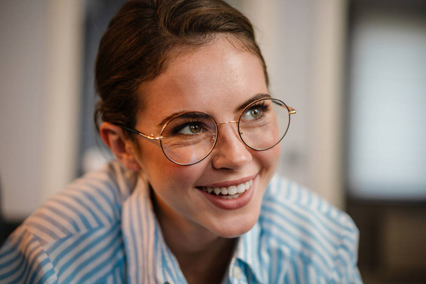 Beautiful happy girl in eyeglasses smiling and looking aside indoors - Foto, Bild