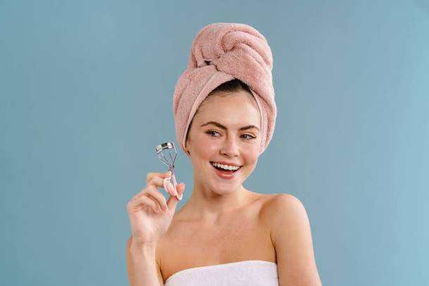 Nice happy girl wearing towels posing with eyelashes curler isolated over blue background - Photo, image