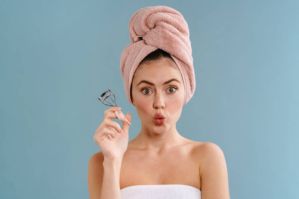 Nice surprised girl wearing towels posing with eyelashes curler isolated over blue background - Foto, afbeelding