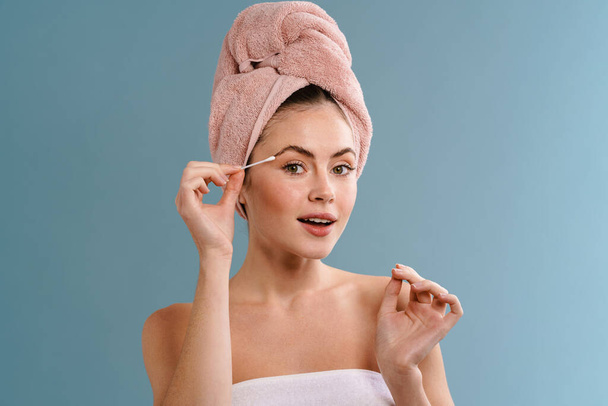 Nice beautiful girl wearing towels doing makeup with cotton swab isolated over blue background - Fotoğraf, Görsel
