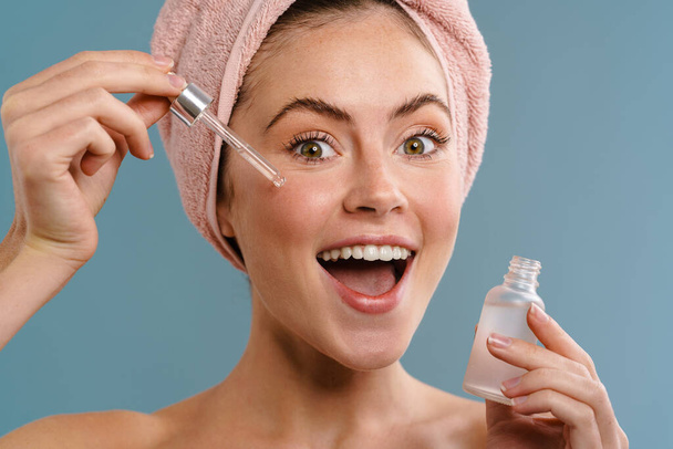 Image of a young cheery optimistic woman in towel taking care of her skin with serum isolated over blue wall background - Foto, immagini