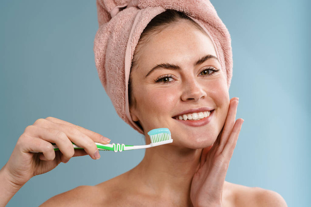 Shirtless happy girl in towel holding toothbrush with toothpaste isolated over blue background - Fotografie, Obrázek