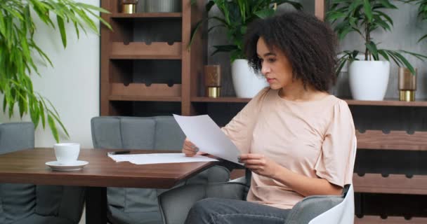 Concentrado mulher afro-americano estudante freelancer com trabalho de papel sentado à mesa no restaurante sala de escritório detém folhas brancas de papel em suas mãos documentos aviso lê cartas dadas relatórios - Filmagem, Vídeo
