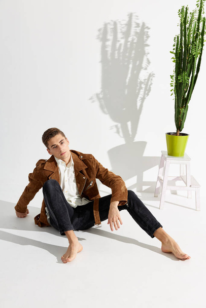A young man in a brown jacket and black trousers sits on the floor near a flower indoors - Φωτογραφία, εικόνα