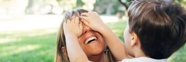 女は子供と遊ぶ。母親の目を覆っている小さな男の子。公園で楽しい時間を過ごしているアル・スモール・キッズの首長たち。母親の概念。母の日。バナー、スライダー3:1パーセント. - 写真・画像
