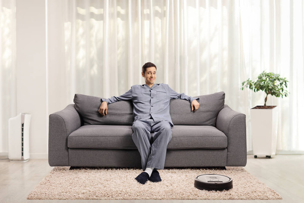 Man in pajamas resting on a sofa at home with a robotic vacuum cleaner on the carpet  - Φωτογραφία, εικόνα