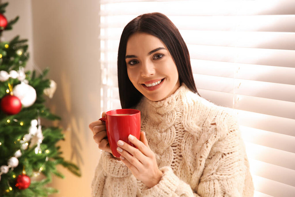 Young woman holding cup of coffee in room with Christmas tree - Foto, Bild