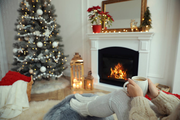 Woman with hot drink resting near fireplace in cozy room decorated for Christmas, closeup - Fotoğraf, Görsel