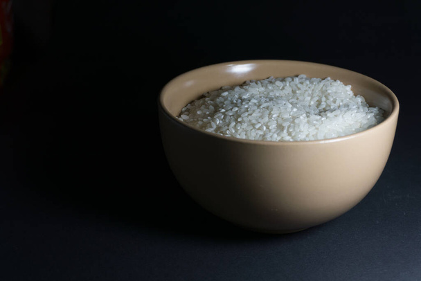 Arroz blanco sin cocer en un tazón beige con granos dispersos cerca sobre un fondo negro - Foto, imagen