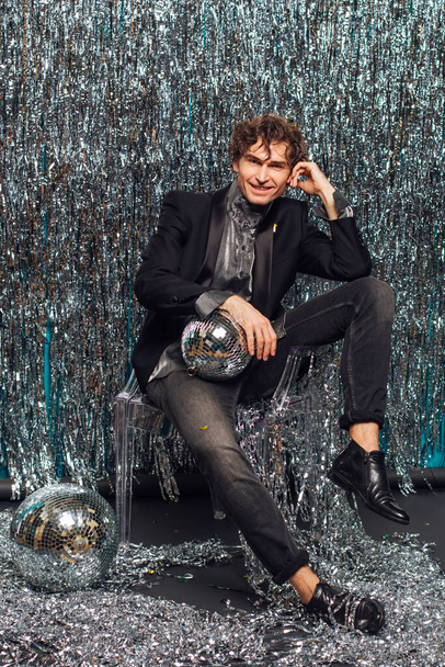 Tall attractive man sitting on the chair over the sparkle background holding mirror disco ball. Party concept. - 写真・画像