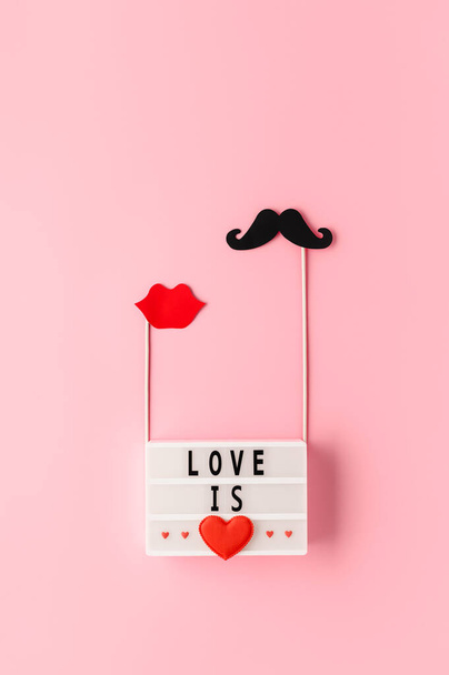 White lightbox with text Love is and paper photo props mustache, lips on pastel pink background. Concept Valentine's Day. Creative flat lay, top view, vertical orientation - Fotoğraf, Görsel