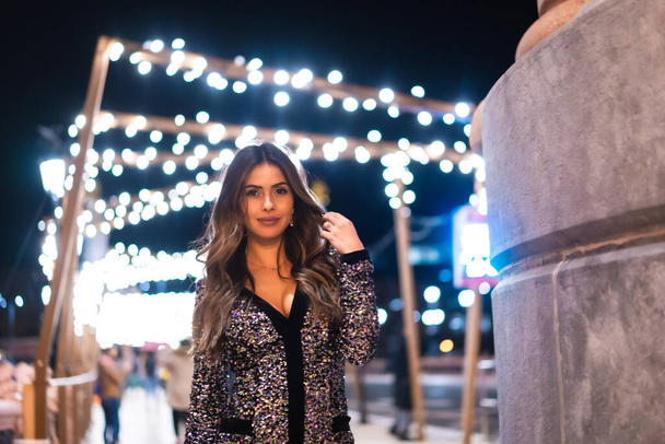 Christmas lifestyle. Portrait a young brunette Caucasian woman in a fashionable dress with sequins, smiling in the background city Christmas lights - Foto, imagen