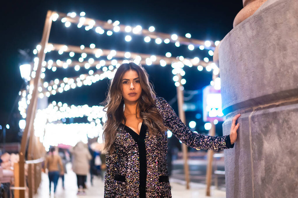 Christmas lifestyle. Portrait a young brunette Caucasian woman in a fashionable dress with sequins, smiling in the background city Christmas lights - Foto, imagen