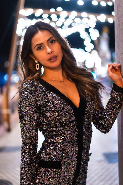 Christmas lifestyle. Portrait a young brunette Caucasian woman in a fashionable dress with sequins, smiling in the Christmas lights of the city, vertical photo - Fotoğraf, Görsel